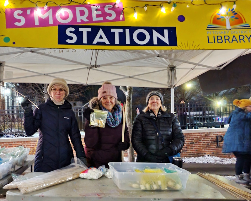 Three women standing at a table outside, under a banner that reads "S'mores Station" with the Petoskey District Library logo